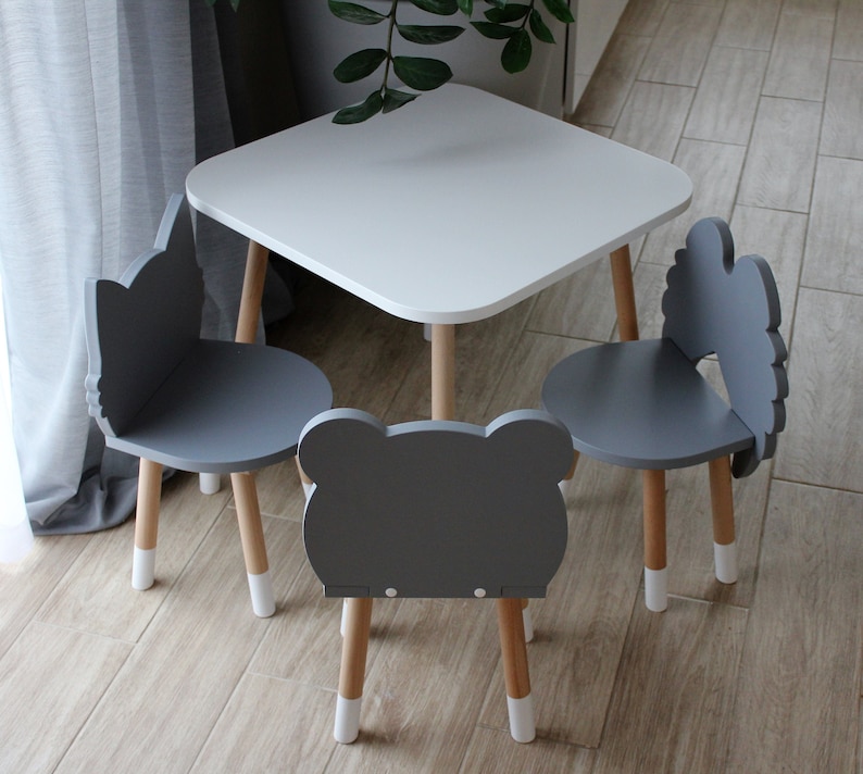 May include: A white square table with rounded edges and three gray chairs. The chairs have animal-shaped backs, including a cat, a bear, and a cloud. The table and chairs are made of wood with white painted legs.