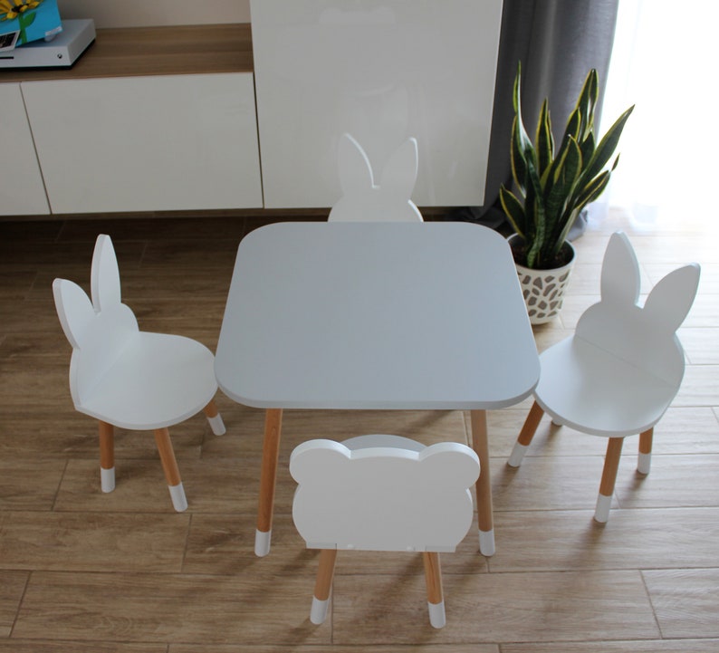 May include: A white table and four white bunny-shaped chairs with wooden legs. The table has a light gray top. The chairs are arranged around the table.