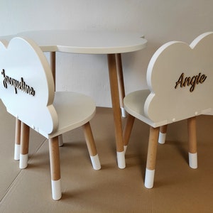 May include: White cloud-shaped children's table and chairs set with wooden legs. The chairs have the names "Jacqueline" and "Angie" written on the backs.