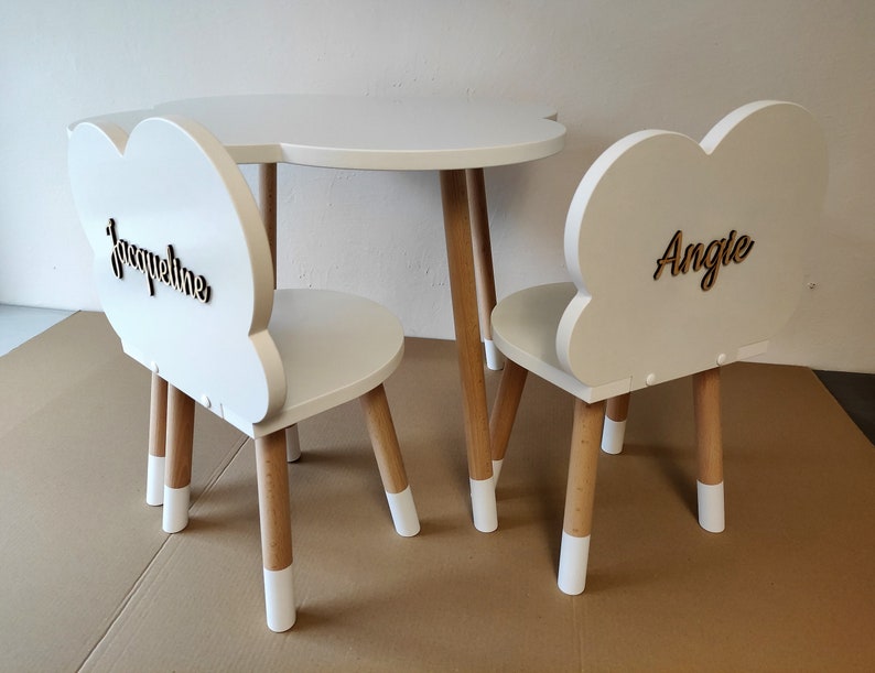 May include: White cloud-shaped children's table and chairs set with wooden legs. The chairs have the names "Jacqueline" and "Angie" written on the backs.