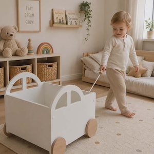 Könnte beinhalten: Ein weißes Holzspielzeugauto mit Holzrädern wird von einem Kind gezogen. Das Auto hat bogenförmige Ausschnitte, die Fenstern ähneln. Die Szene spielt in einem Kinderzimmer mit neutraler Farbpalette, einem Teddybär und einem Schild mit der Aufschrift "PLAY DREAM GROW".