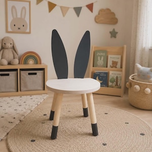 Op de afbeelding: Een kinderstoel met een konijnenoordesign. De stoel heeft een ronde, witte zitting en vier houten poten met zwarte uiteinden. De rugleuning van de stoel heeft twee grote, donkergrijze konijnenoren. De stoel staat in een kinderkamer.