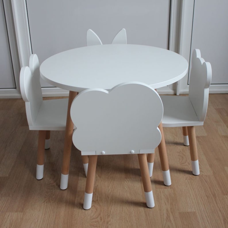 May include: A white children's table and four chairs set. The table is round and the chairs have unique animal-shaped backs. The chairs have wooden legs with white painted tips.