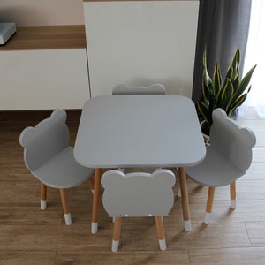 May include: A gray children's table and four chairs with bear-shaped backs. The table and chairs have a light gray finish and wooden legs.