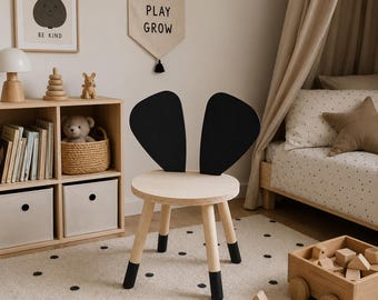 May include: A wooden children's chair with a round seat and black bunny ear-shaped backrest. The chair has four legs with black tips. The room also features a wooden toy wagon, a framed pear print, and a banner that says "DREAM PLAY GROW."