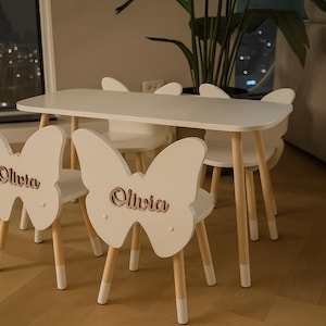 May include: A white children's table and chairs set. The chairs have butterfly-shaped backs with the name "Olivia" cut out. The table and chairs have light-coloured wooden legs. The set is designed for children.