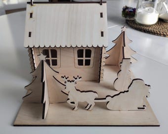 Può includere: Una scena di villaggio natalizio in legno con una casa, alberi, una renna e una slitta. La casa ha un tetto bianco e pareti marroni con finestre. Gli alberi sono verdi e la renna è marrone. La slitta è vuota.