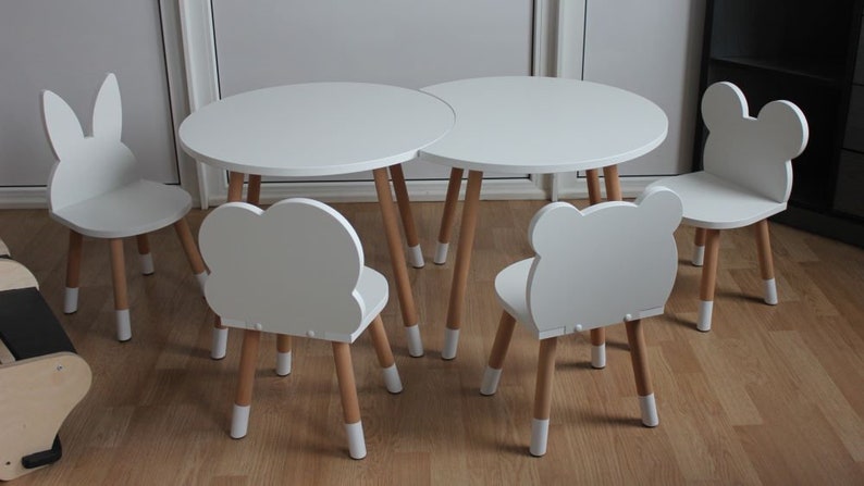 May include: A white children's table and four chairs. The table is round and has a white top and wooden legs. The chairs are all different shapes, with animal-shaped backs. The chairs have white tops and wooden legs.