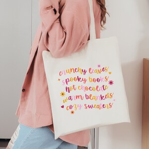 May include: A white canvas tote bag with the text "crunchy leaves, spooky books, hot chocolate, warm blankets, cozy sweaters" printed in pink, orange, and yellow with small flower and heart accents.