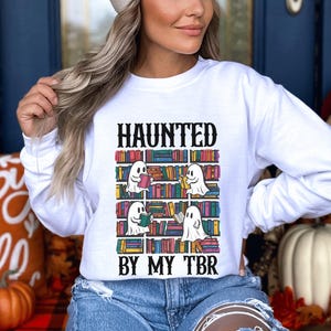May include: White sweatshirt with a graphic of ghosts reading books on a bookshelf. The text "Haunted by my TBR" is printed below the graphic.