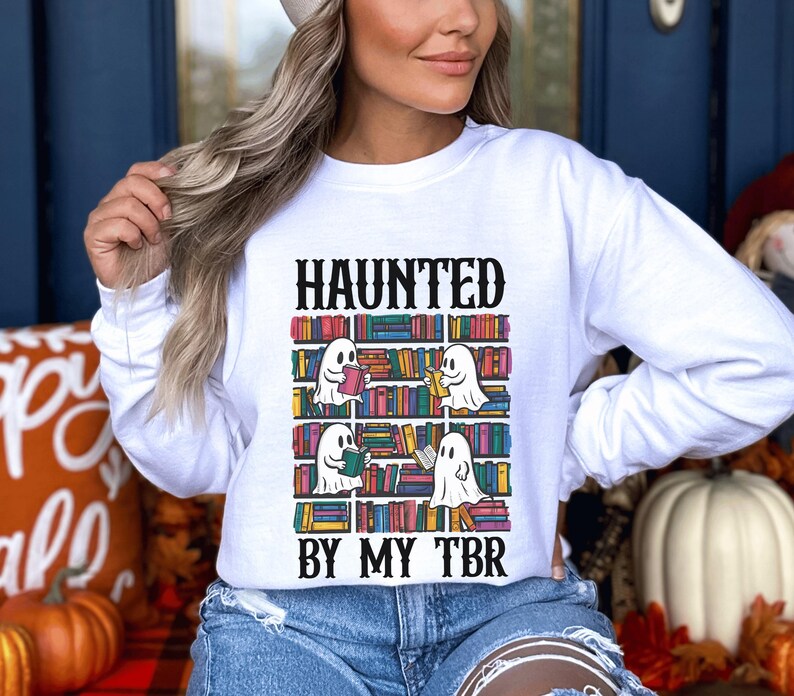 May include: White sweatshirt with a graphic of ghosts reading books on a bookshelf. The text "Haunted by my TBR" is printed below the graphic.