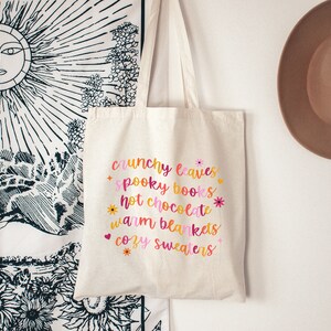 May include: A white canvas tote bag with the text "crunchy leaves, spooky books, hot chocolate, warm blankets, cozy sweaters" printed in pink, orange, and yellow with floral accents.