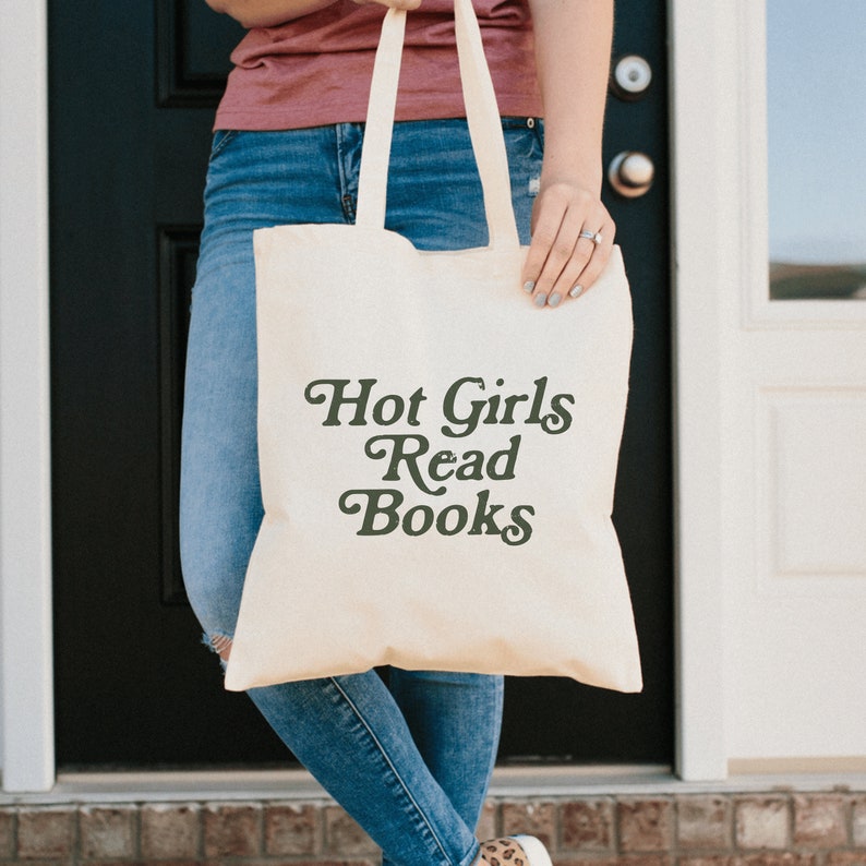 May include: Cream-colored tote bag with the text "Hot Girls Read Books" in a dark green, stylized font. The bag has long, matching handles and is held by a person wearing blue jeans and a maroon shirt.