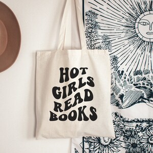K&ouml;nnte beinhalten: Eine wei&szlig;e Canvas-Tragetasche mit schwarzem Text, der "HOT GIRLS READ BOOKS" in einer Retro-Schriftart lautet.