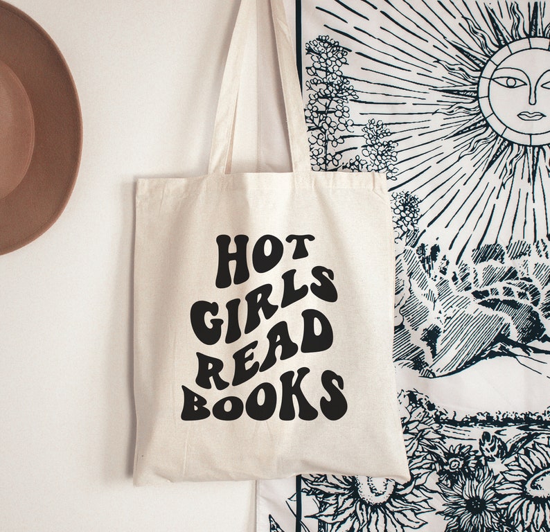 K&ouml;nnte beinhalten: Eine wei&szlig;e Canvas-Tragetasche mit schwarzem Text, der "HOT GIRLS READ BOOKS" in einer Retro-Schriftart lautet.