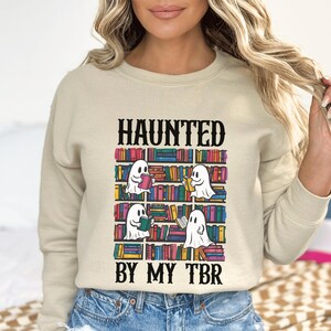 May include: A beige sweatshirt with the text "HAUNTED BY MY TBR" printed on the front. The text is in black and the graphic features a bookshelf with ghosts reading books.