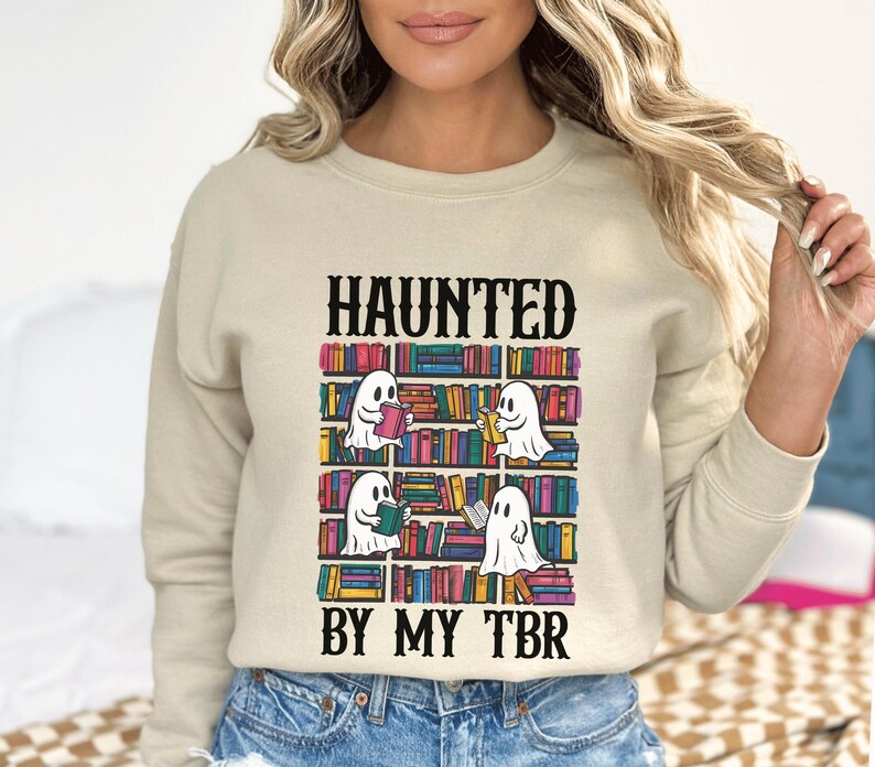 May include: A beige sweatshirt with the text "HAUNTED BY MY TBR" printed on the front. The text is in black and the graphic features a bookshelf with ghosts reading books.