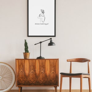 May include: A black framed print with a line drawing of a person with flowers on their head. The text "obstinate, headstrong girl" is below the drawing.