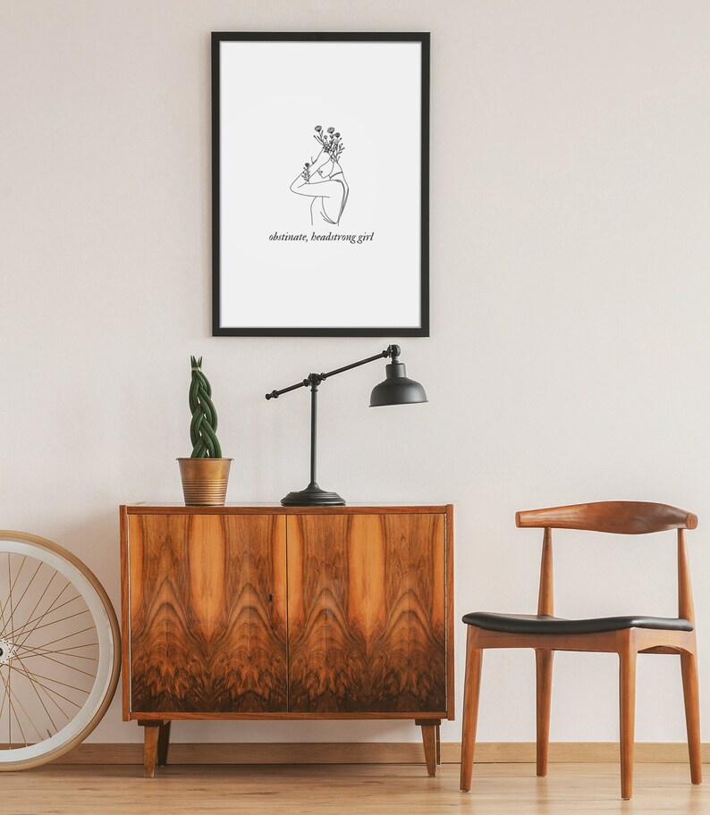 May include: A black framed print with a line drawing of a person with flowers on their head. The text "obstinate, headstrong girl" is below the drawing.