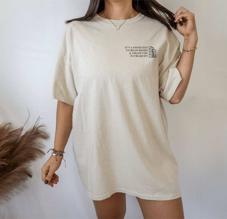 Pode incluir: Uma camiseta bege oversized com o texto "It's a good day to read books & smash the patriarchy" estampado na frente.