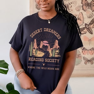 Op de afbeelding: Zwart T-shirt met een afbeelding van een woestijnlandschap met de tekst "Desert Dreamers Reading Society Where the Wild Books Are"