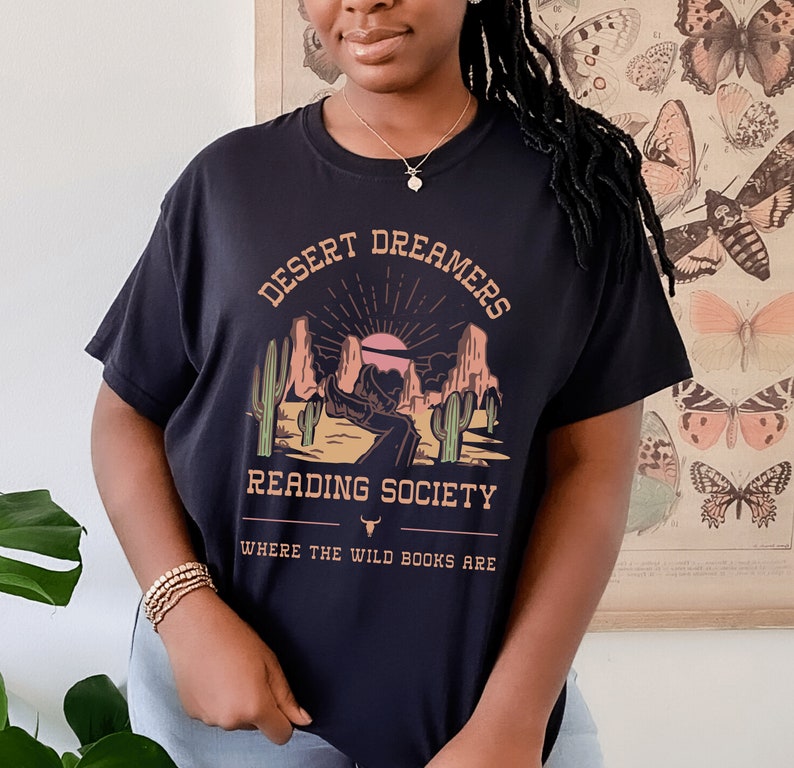 Op de afbeelding: Zwart T-shirt met een afbeelding van een woestijnlandschap met de tekst "Desert Dreamers Reading Society Where the Wild Books Are"