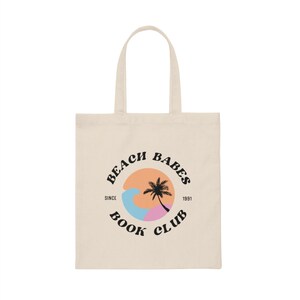 以下が含まれることがあります： ベージュのトートバッグに、黒い円のロゴがあり、「Beach Babes Book Club」と書いてあり、円の中にヤシの木と波があります。「Since 1991」という文字が円のすぐ下にあります。