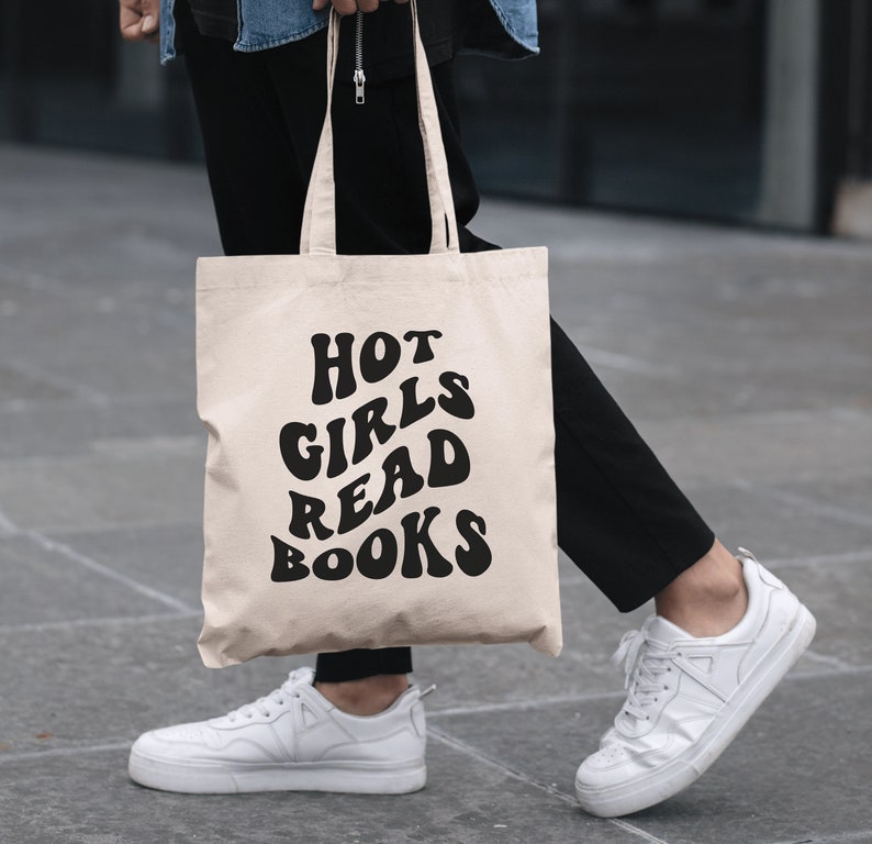 K&ouml;nnte beinhalten: Eine beige Tragetasche mit schwarzem Text, der in einer Retro-Schrift "HOT GIRLS READ BOOKS" lautet.