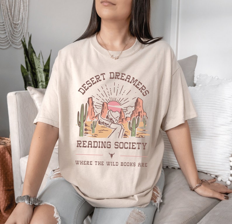 Op de afbeelding: Een beige T-shirt met een grafische print van een woestijnlandschap met zonsondergang, cactussen en de tekst "Desert Dreamers Reading Society Where the Wild Books Are".