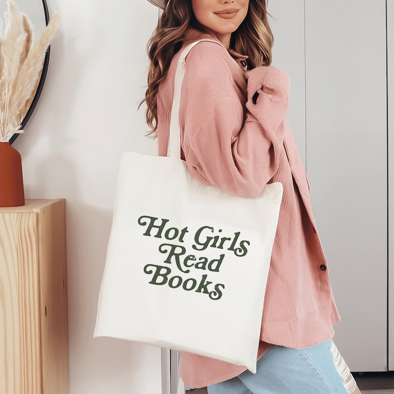 May include: Cream-colored tote bag with the text "Hot Girls Read Books" in dark green script. The bag has long handles and is made of a sturdy fabric. The tote bag is a stylish accessory for carrying books and other items.