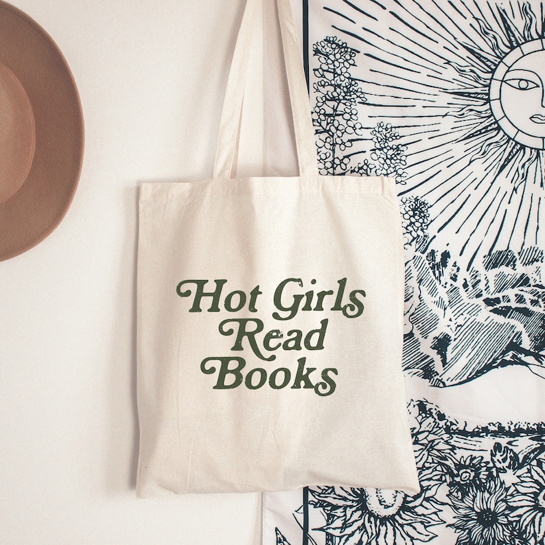 May include: A cream-colored tote bag with the text "Hot Girls Read Books" in dark green script. The bag is hanging on a white wall, with a brown hat and a patterned tapestry in the background. The tote bag has long handles.