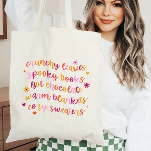May include: A white tote bag with the text "crunchy leaves", "spooky books", "hot chocolate", "warm blankets", and "cozy sweaters" printed in pink, orange, and yellow.