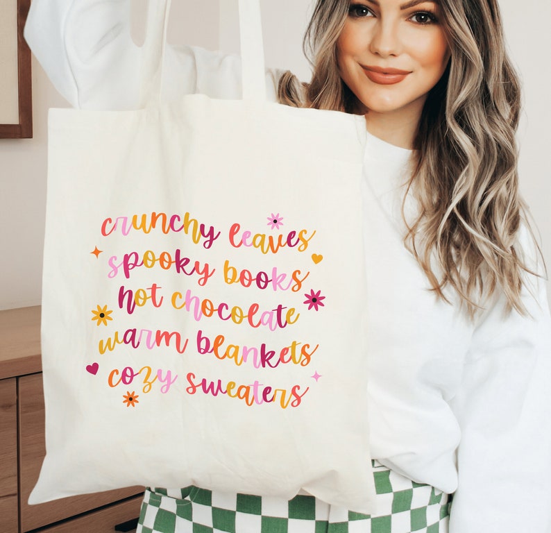 May include: A white tote bag with the text "crunchy leaves", "spooky books", "hot chocolate", "warm blankets", and "cozy sweaters" printed in pink, orange, and yellow.