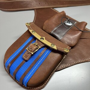 K&ouml;nnte beinhalten: Ein brauner Lederg&uuml;rtel mit zwei befestigten Taschen. Die Haupttasche hat einen leuchtend blauen Stoffstreifen, einen Messingschnallenverschluss und Messingnieten. Eine kleinere Zusatztasche ist mit einem dunklen Lederriemen und Knopf gesichert.