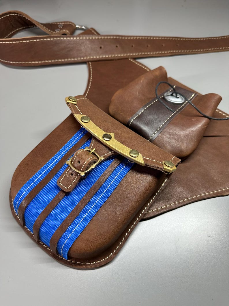 K&ouml;nnte beinhalten: Ein brauner Lederg&uuml;rtel mit zwei befestigten Taschen. Die Haupttasche hat einen leuchtend blauen Stoffstreifen, einen Messingschnallenverschluss und Messingnieten. Eine kleinere Zusatztasche ist mit einem dunklen Lederriemen und Knopf gesichert.