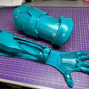 K&ouml;nnte beinhalten: Ein Paar t&uuml;rkisfarbene Stulpenhandschuhe mit einem segmentierten Unterarmteil mit geometrischen Ausschnitten und einem passenden Handschuh mit beweglichen Fingerplatten, die an einem Stoff&auml;rmel befestigt sind, f&uuml;r Schutz oder Kost&uuml;me.