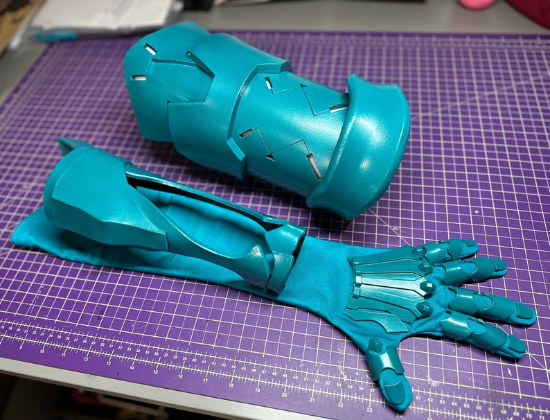 K&ouml;nnte beinhalten: Ein Paar t&uuml;rkisfarbene Stulpenhandschuhe mit einem segmentierten Unterarmteil mit geometrischen Ausschnitten und einem passenden Handschuh mit beweglichen Fingerplatten, die an einem Stoff&auml;rmel befestigt sind, f&uuml;r Schutz oder Kost&uuml;me.