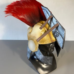 K&ouml;nnte beinhalten: Ein Helm mit einer verwitterten cremefarbenen Kuppel, einem gro&szlig;en leuchtend roten Kamm und metallisch silberfarbenen Seitenplatten. Das Design umfasst eine markante, kantige silberne Visierverl&auml;ngerung und dunkelbraune St&uuml;tzstangen vorne.