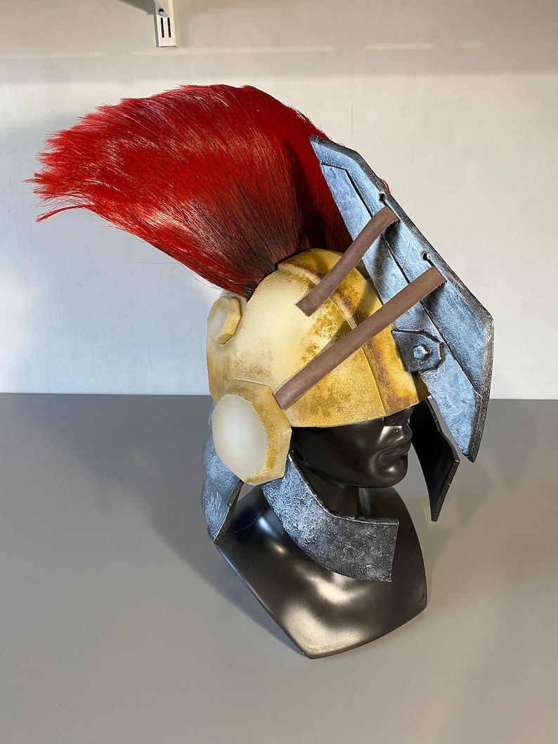 K&ouml;nnte beinhalten: Ein Helm mit einer verwitterten cremefarbenen Kuppel, einem gro&szlig;en leuchtend roten Kamm und metallisch silberfarbenen Seitenplatten. Das Design umfasst eine markante, kantige silberne Visierverl&auml;ngerung und dunkelbraune St&uuml;tzstangen vorne.