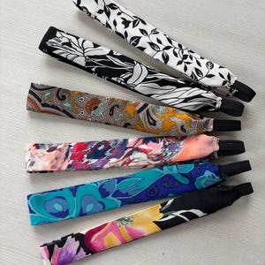 May include: Six colorful patterned headbands with elastic bands. The headbands feature floral and paisley designs in shades of black, white, blue, red, orange, and yellow.