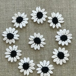 May include: A collection of ten white daisy flower appliques with black centers arranged on a textured, light gray surface. The appliques are made of a delicate, lace-like material, perfect for embellishing clothing or crafts.