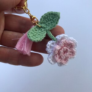 May include: A pink and white crocheted flower keychain with green crocheted leaves and a pink tassel.