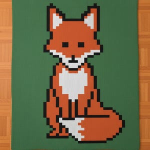 Puede incluir: Una manta de punto con un diseño de zorro pixelado. El zorro es principalmente naranja con detalles blancos y negros, sobre un fondo verde. La manta es rectangular y parece estar hecha de material suave.