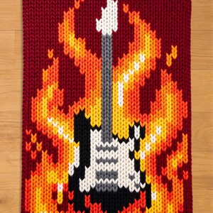 May include: A knitted guitar design with flames in shades of yellow, orange, and red. The guitar is black, white, and gray, set against a burgundy background. The image is a close-up of a knitted item.