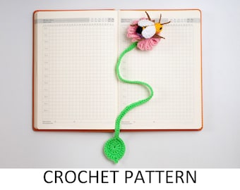 May include: A green crocheted flower bookmark with a pink flower and a yellow and black bee on top. The bookmark is attached to a notebook.