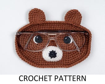 May include: Crocheted brown bear-shaped glasses case with a white nose and black eyes. The case is holding a pair of brown-framed glasses.