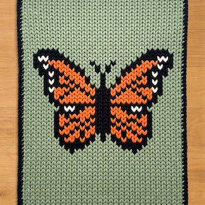 May include: A knitted square rug featuring a butterfly design. The butterfly has orange and black wings with white accents, set against a light green background. The rug has a dark blue border.