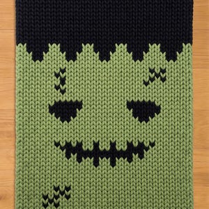Könnte beinhalten: Eine rechteckige Strickdecke mit einem grünen Gesicht, schwarzem Haar, schwarzen Augen, einem gezackten, schwarz gestickten Mund sowie schwarz gestickten Narben auf der Stirn und der unteren Wange. Das Strickmuster ist durchgehend dick.