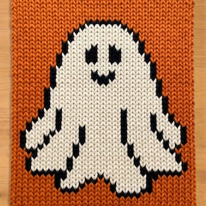 Könnte beinhalten: Eine rechteckige Strickdecke mit einem großen weißen Geist mit schwarzen Augen und einem schwarzen Mund, zentriert auf einem einfarbigen orangefarbenen Hintergrund. Das Strickmuster ist grob und gleichmäßig, mit schwarzem Garn umrandet.