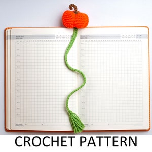 May include: A green crocheted pumpkin bookmark with a brown stem and orange crocheted pumpkin. The bookmark is laying on a closed orange and white gridded notebook.